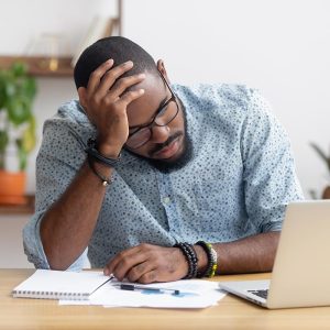 Man holding his head looking at computer and paperwork